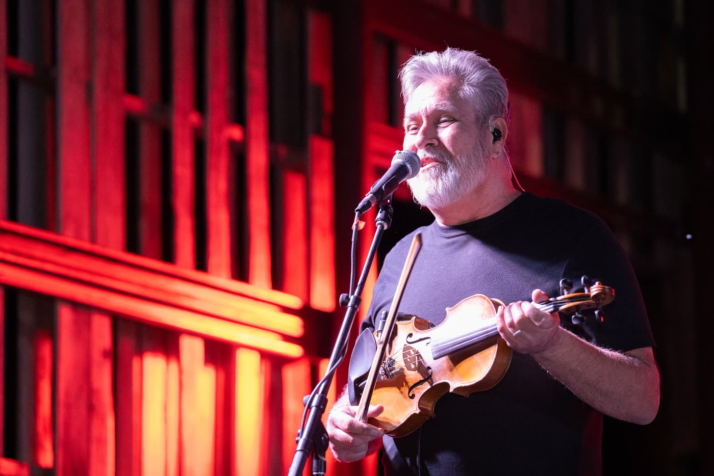 A fiddle player lit in deep red against the pallet-wood back wall of the Coyote stage.