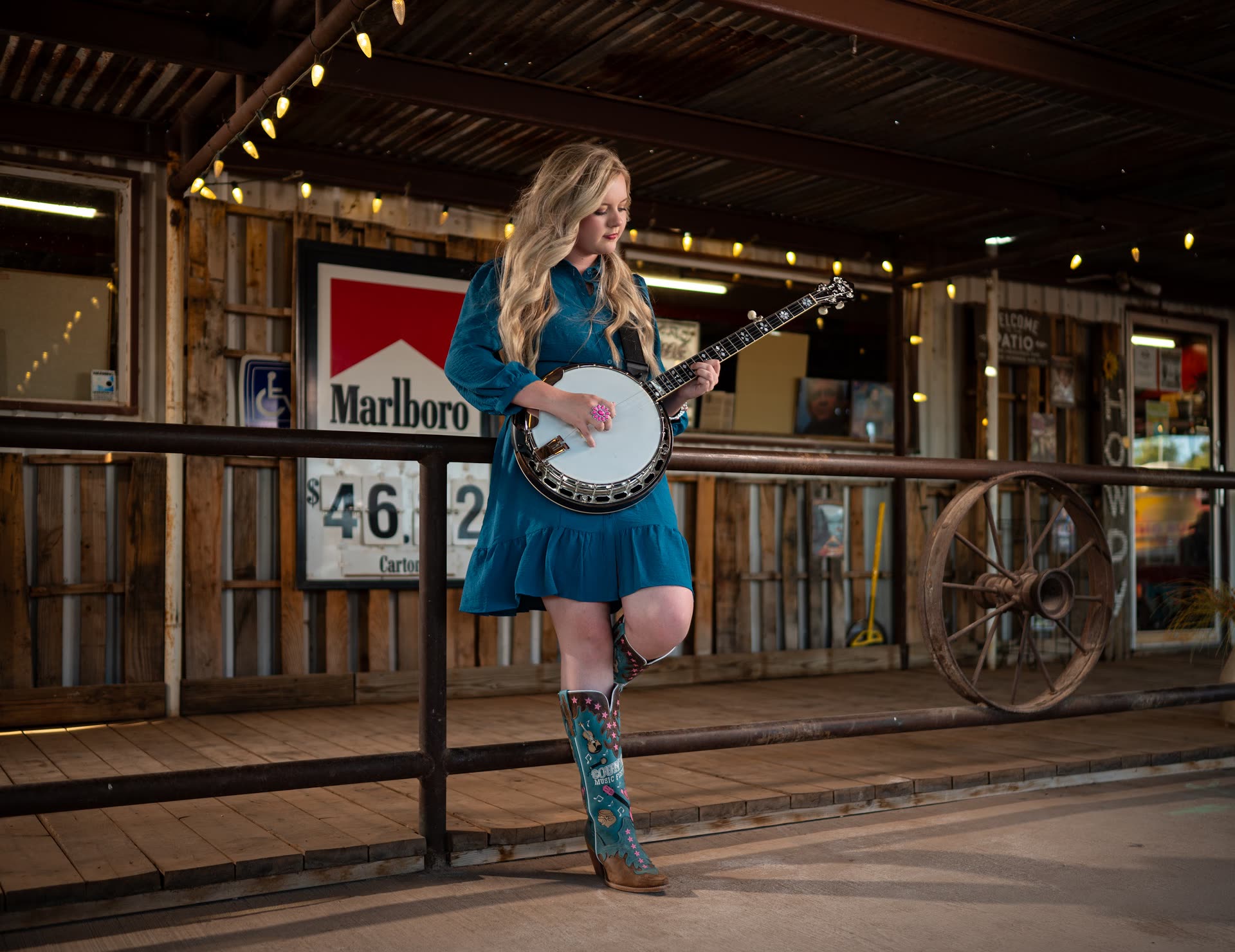 Hadlie Jo playing banjo on the front porch of The Coyote Store, leaning on the rail beneath the string lights, with the Marlboro sign, the carton price board and an old wagon wheel behind her.