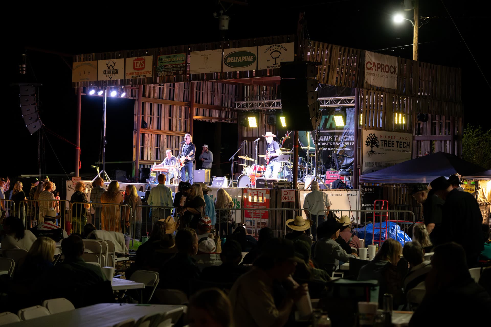 A night concert at The Coyote Store in Gail, Texas: the pallet-wood stage lit up, sponsor banners overhead, and a full crowd seated at tables under the open sky.